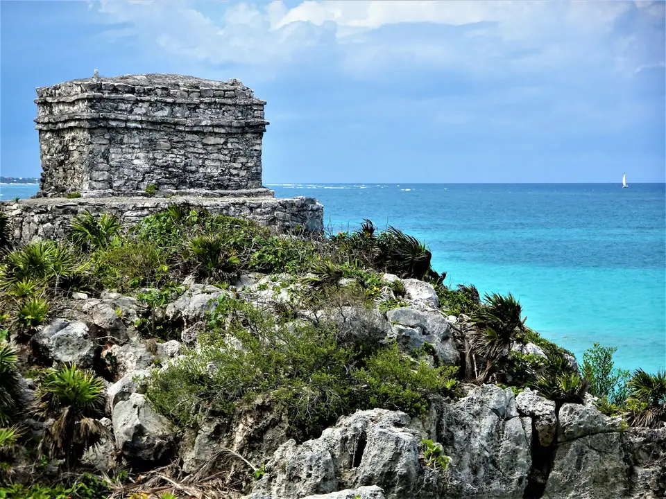 a stone structure on a rocky cliff overlooking the ocean with Tulum in the background — Tulum